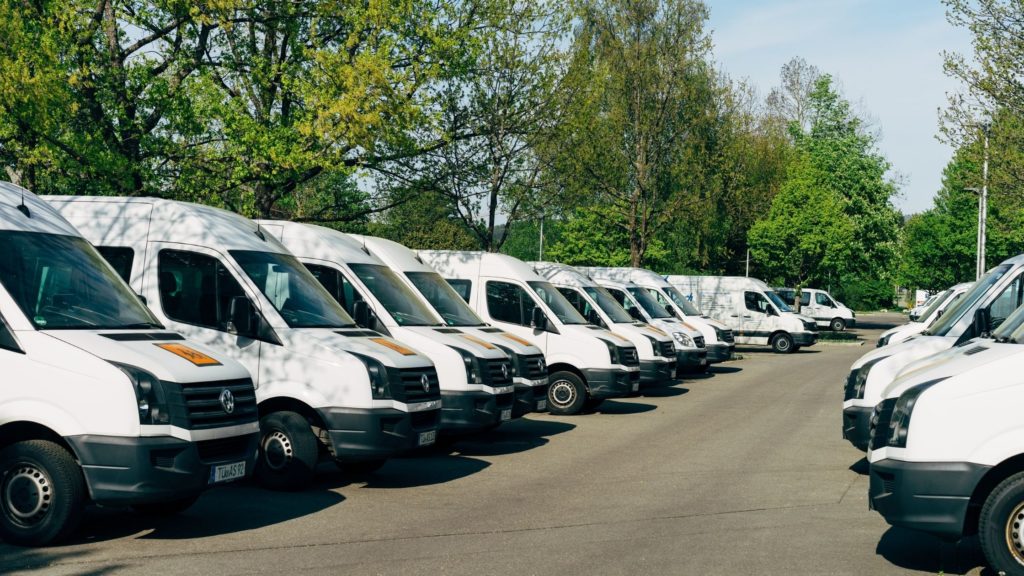 Rows of white vans parked up by some trees.